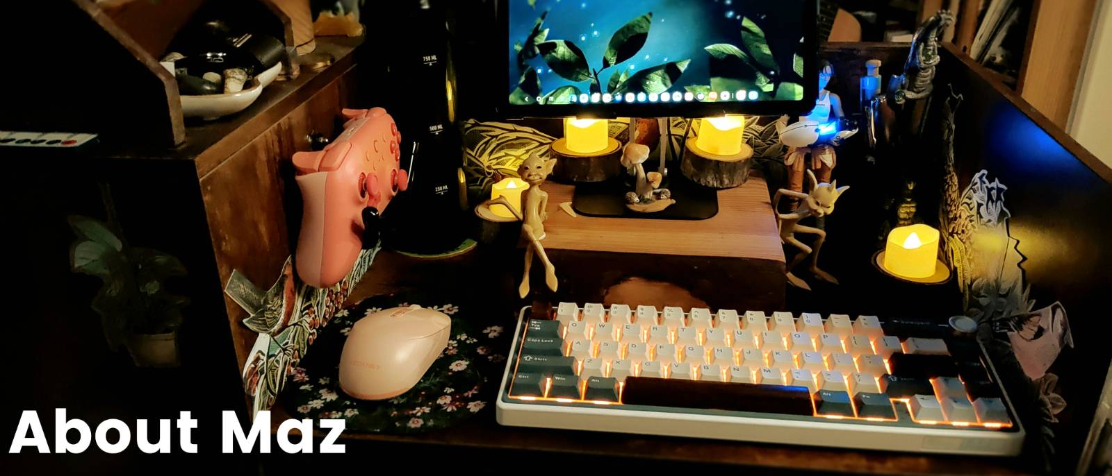 A photograph of a desk that is full of desk tools and ephemera. A screen, a mmouse and a keyboard are all visible, but there are also a number of lit candles, a game controller, some figures of pixies created by Wendy Froud and a figure of Chell from the Portal games. There is also a water bottle and some decorations of birds attached to the sides of the desk area. The words About Maz are shown in the bottom left of the image.