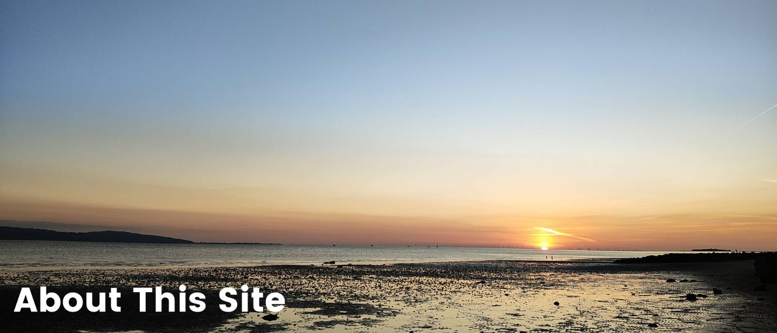 A photograph of a very late sunset on a beach. The sky is glowing with the gradient of light and the incoming tight is lit up on the beach. The sun is just on the horizon and is very yellow. The words About This Site appear in the bottom left of the image.