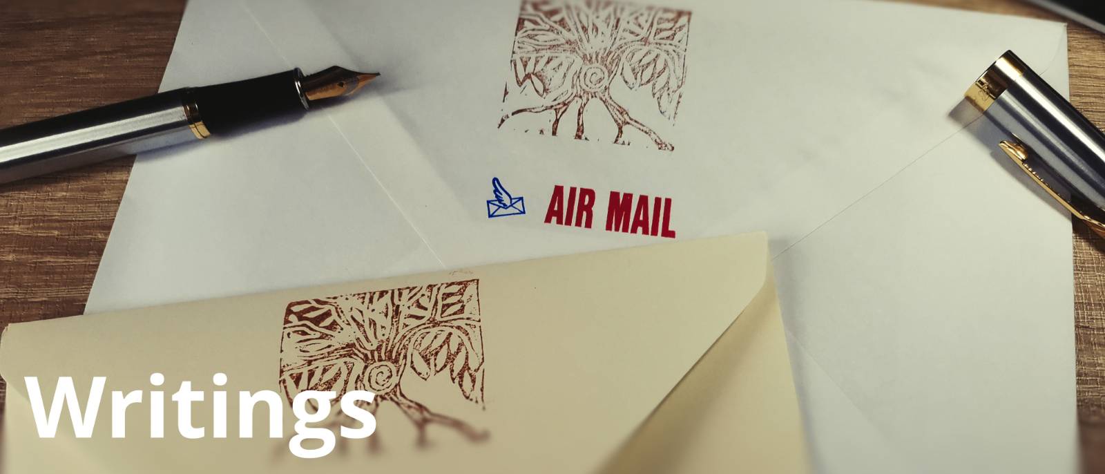 A photograph of a envelopes and a fountain pen on a desk. The envelopes have printed designs of a tree on the flap and the words Air Mail appear on one of the. The cap is off of the fountain pen. The word Writings appears in the bottom left of the image.